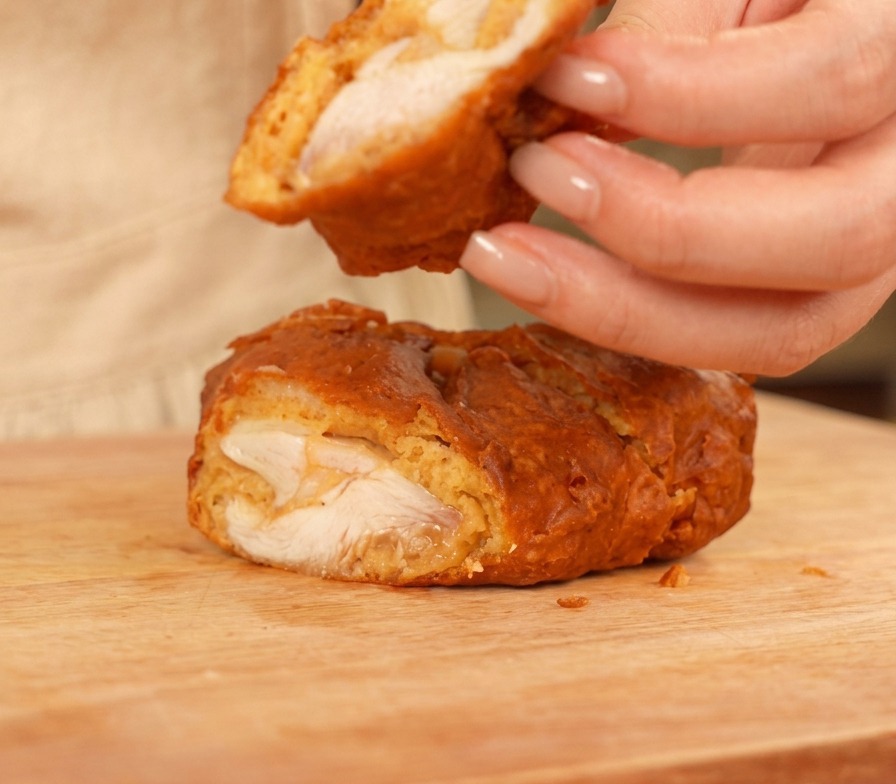 A sliced piece of fried chicken on a wooden cutting board, showing the white juicy meat and a thick, crunchy golden crust.