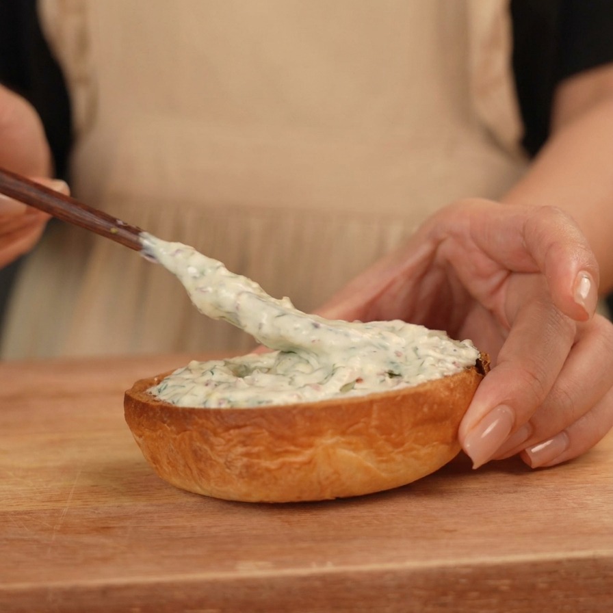 A hand using a small wooden spreader to apply a thick, creamy white sauce onto the bottom half of a toasted burger bun.