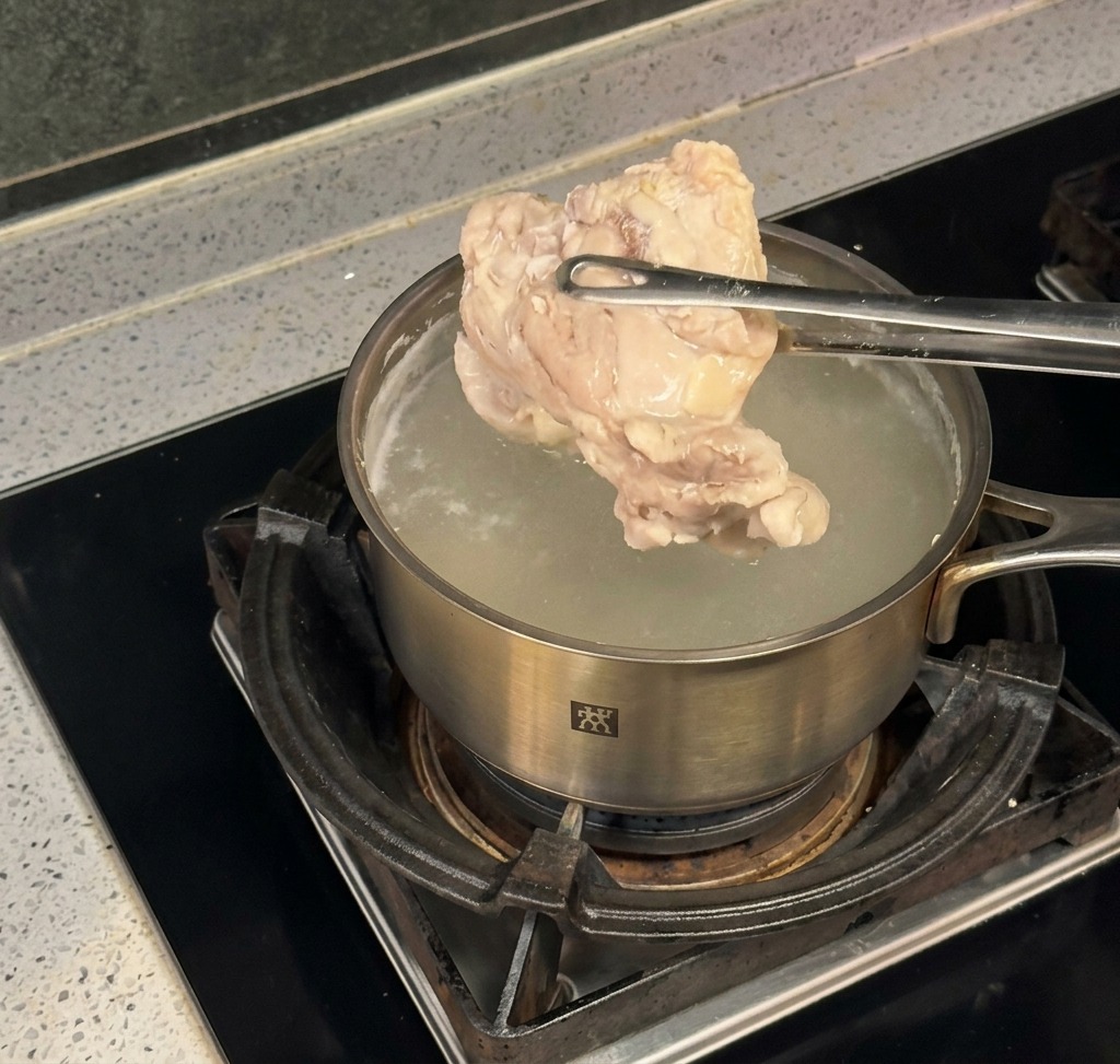 A pair of metal tongs lifting a large piece of steamed, cooked chicken out of a stainless steel pot filled with boiling water on a stove.