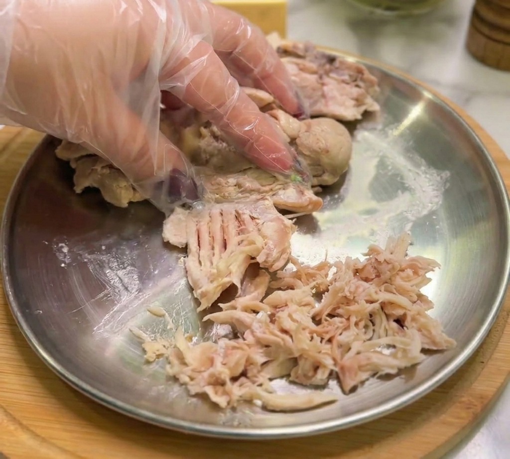 A gloved hand pulling apart cooked chicken into thin, shredded strips on a flat metal serving plate.