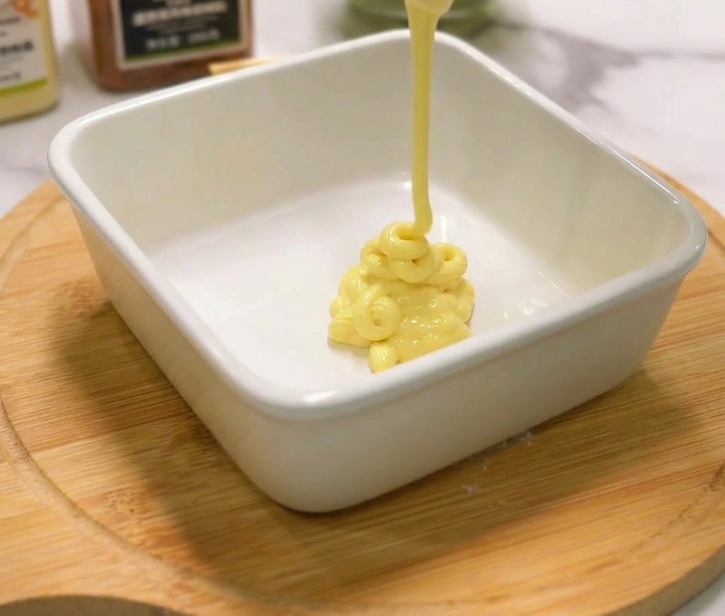 Creamy yellow mayonnaise being squeezed from a bottle into a small white square ceramic bowl on a wooden board.