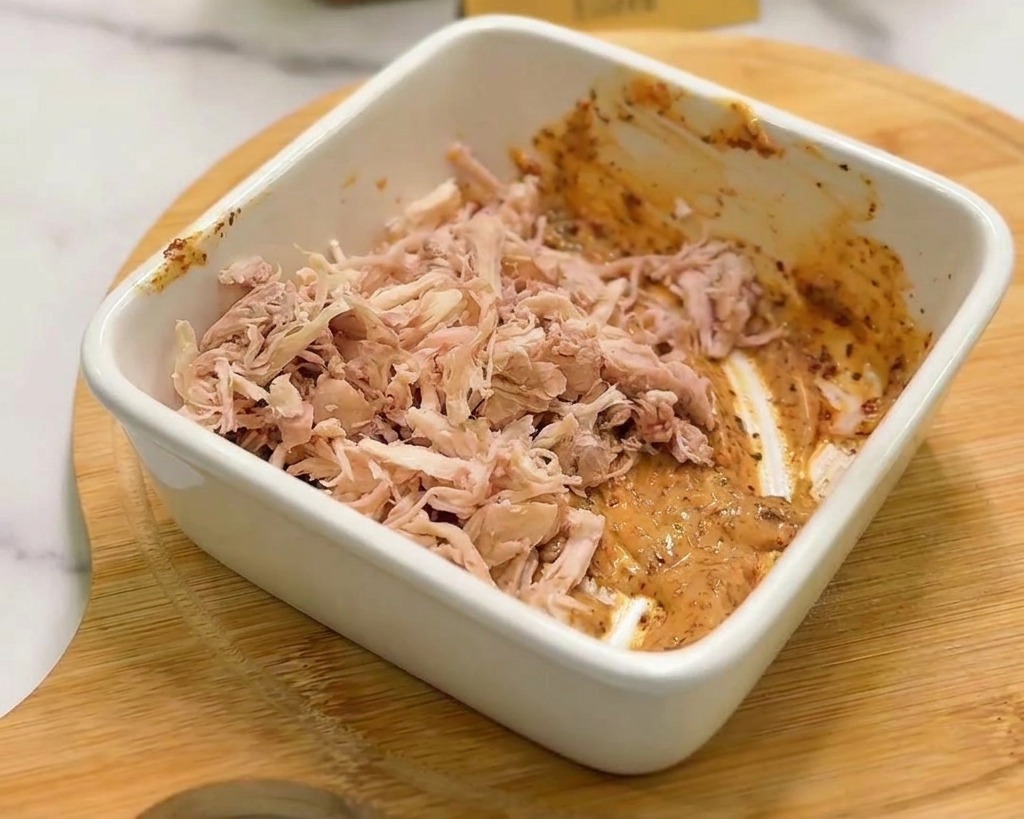 A pile of shredded cooked chicken being mixed into a creamy orange spicy sauce inside a white square ceramic bowl.