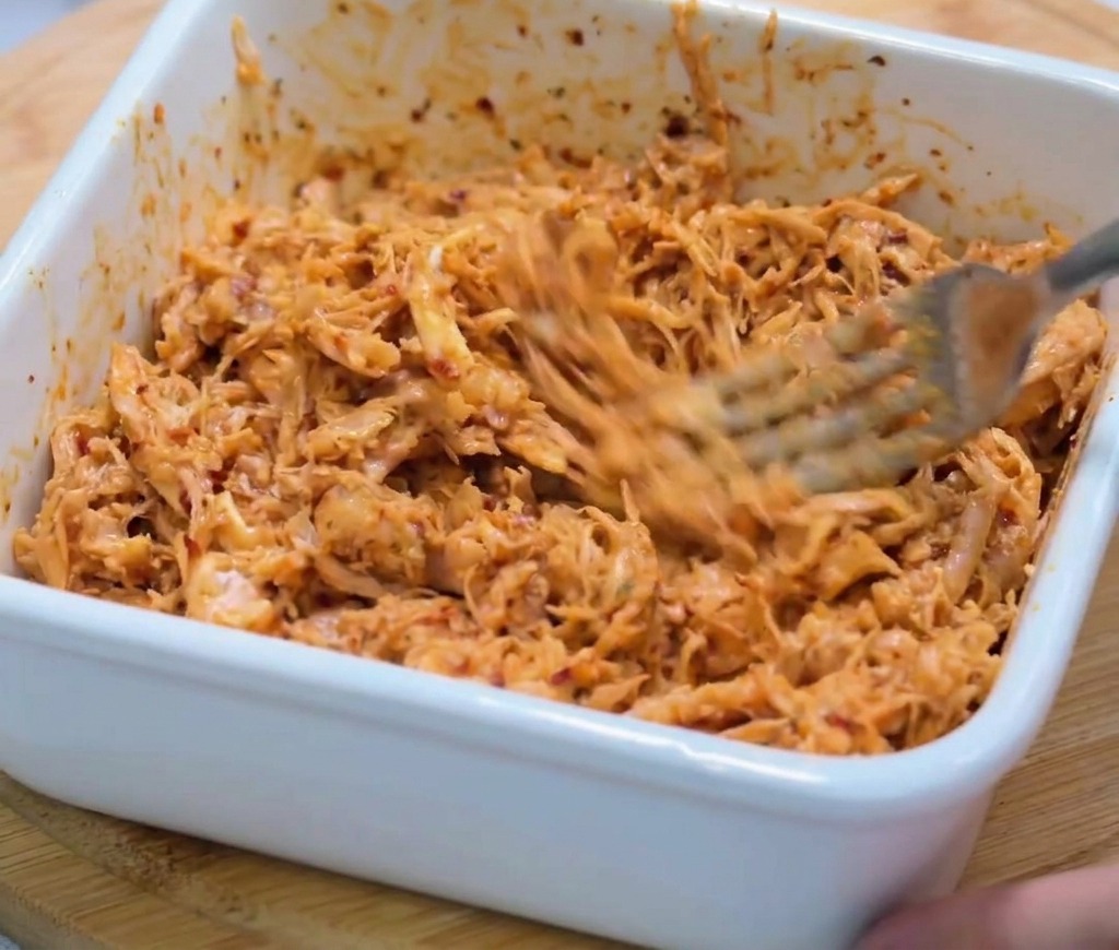 A close-up shot of finely shredded chicken being stirred into a thick, spicy red sauce within a white square ceramic bowl using a metal fork.
