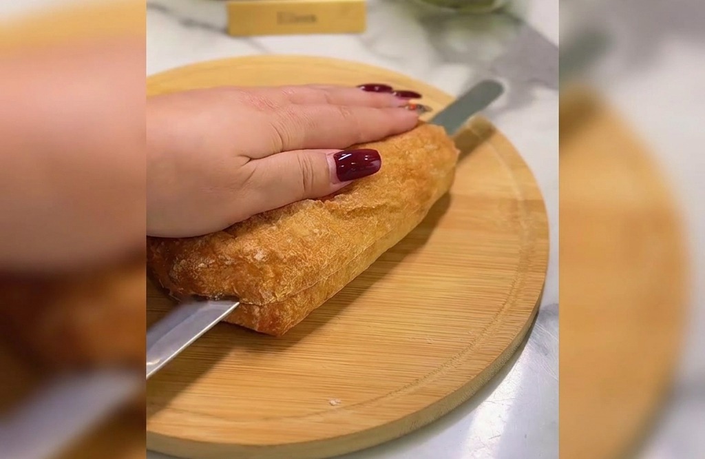 A hand holding a golden ciabatta loaf while a serrated bread knife slices through the middle on a round wooden board.