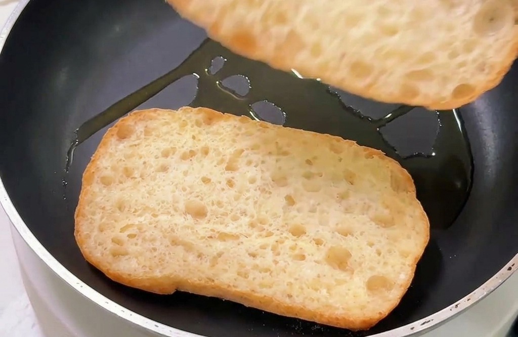 Two pieces of ciabatta bread being toasted cut-side down in a non-stick skillet with a light coating of oil.