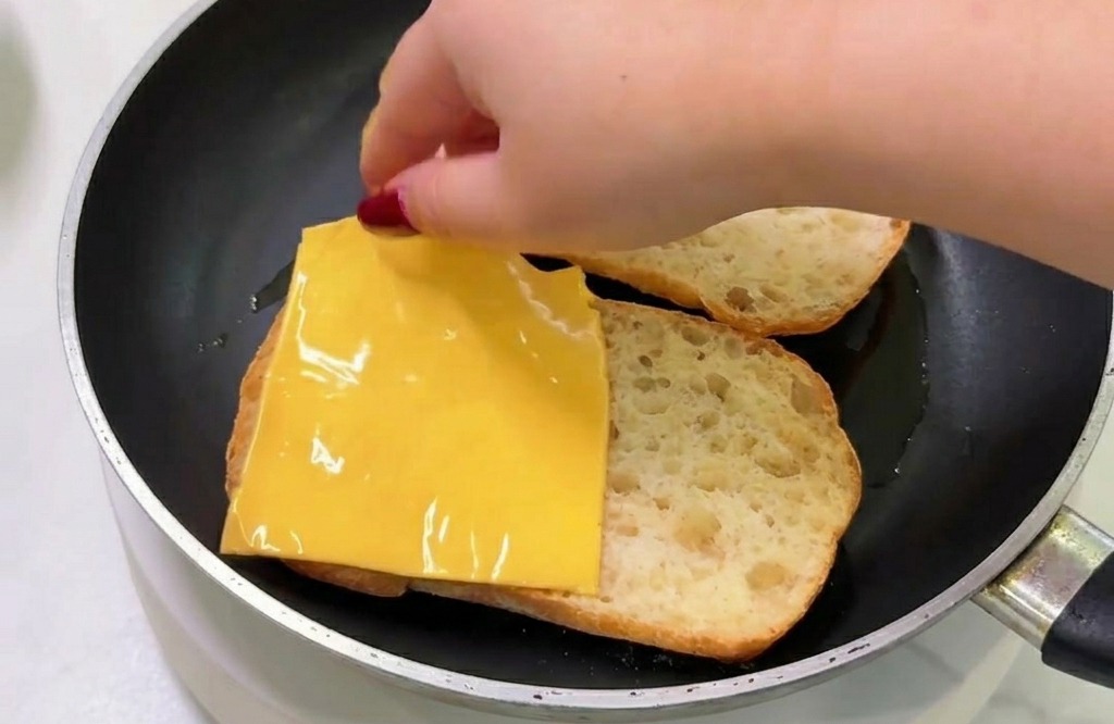 A hand placing a square slice of yellow processed cheese onto a piece of toasted bread inside a frying pan.