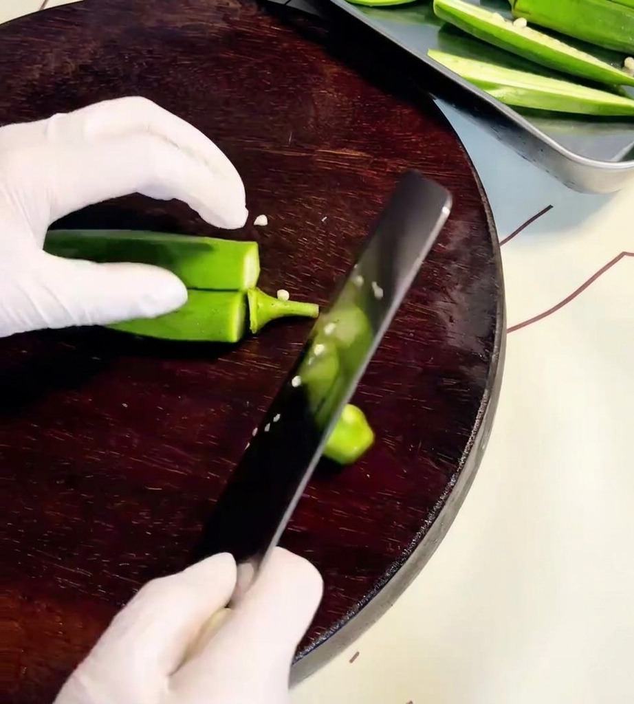 A close-up view of hands using a knife to slice okra pods open on a wooden cutting board.