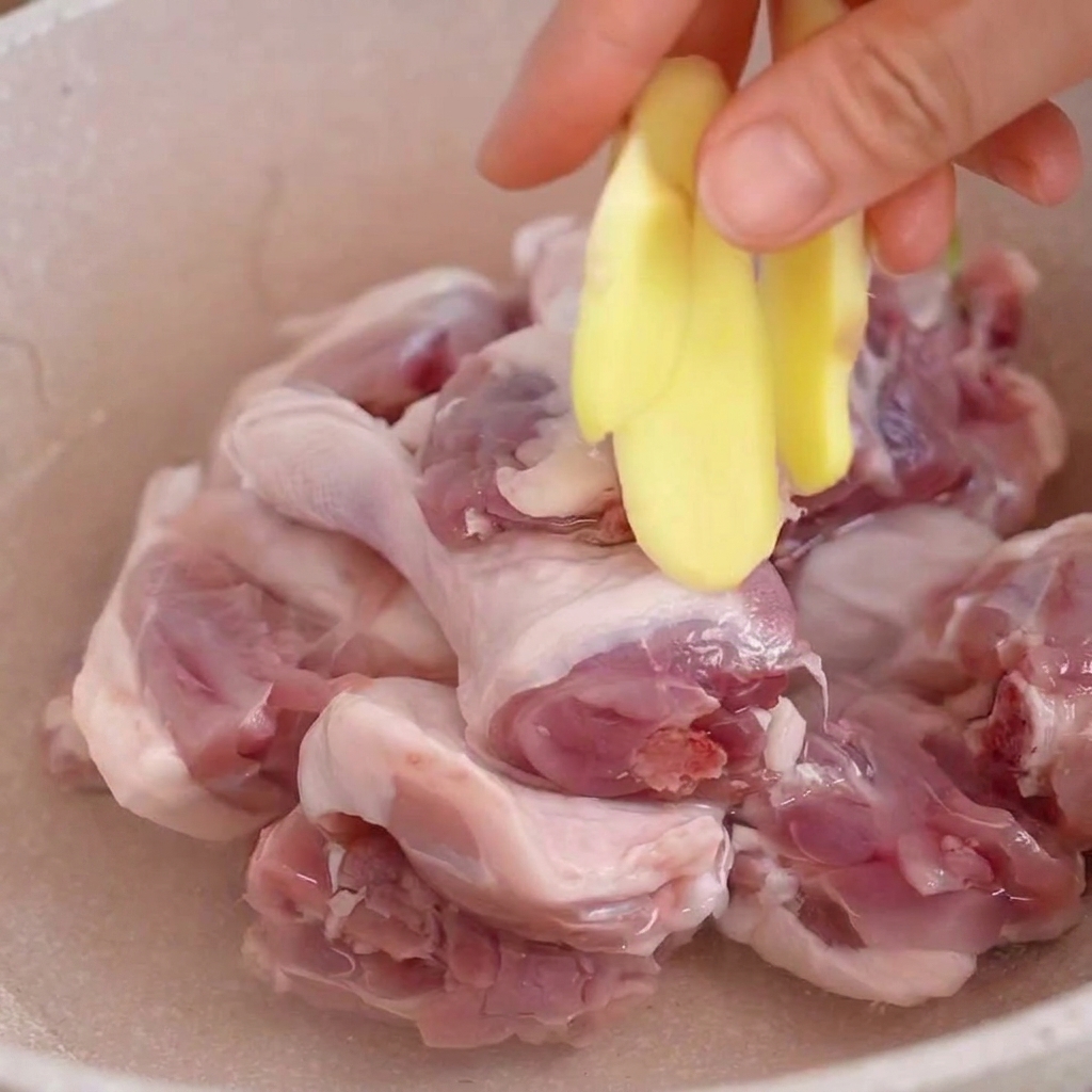 A hand adding fresh ginger slices into a pot with raw duck pieces submerged in water.