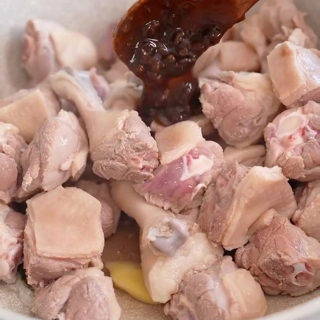 A wooden spoon scooping dark bean paste onto pieces of duck meat cooking in a light-colored pan.