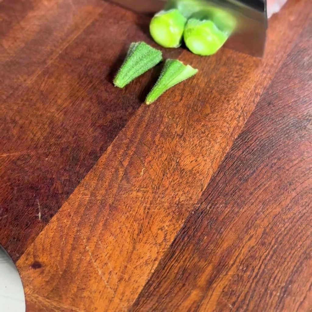 A close-up view of blanched okra pods on a wooden cutting board with the stem ends being sliced off.