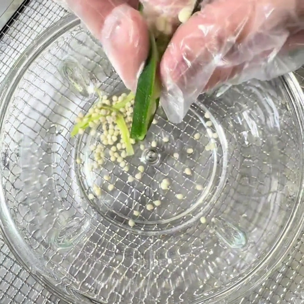 Okra seeds being scraped out of the halved pods into a clear glass bowl.