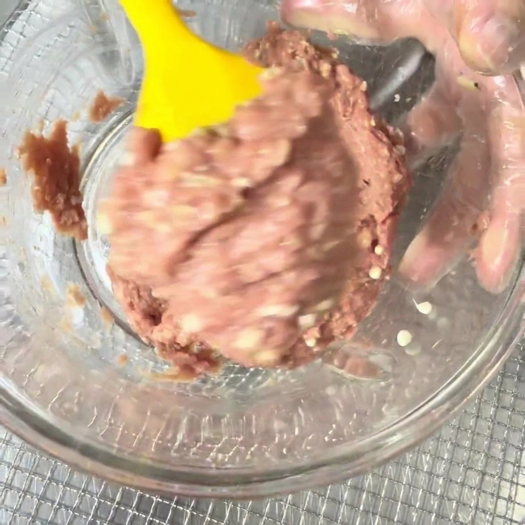 Minced duck meat and okra seeds being combined in a bowl with a yellow silicone spatula.
