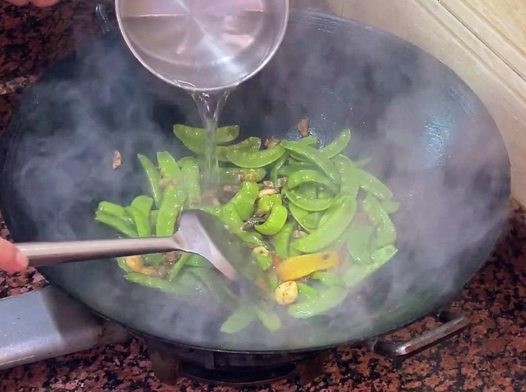 Warm water being poured into the wok with the stir-frying snow peas and sausage.