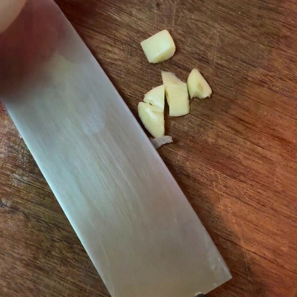 Pieces of roughly chopped garlic cloves next to a large metal knife on a wooden cutting surface.