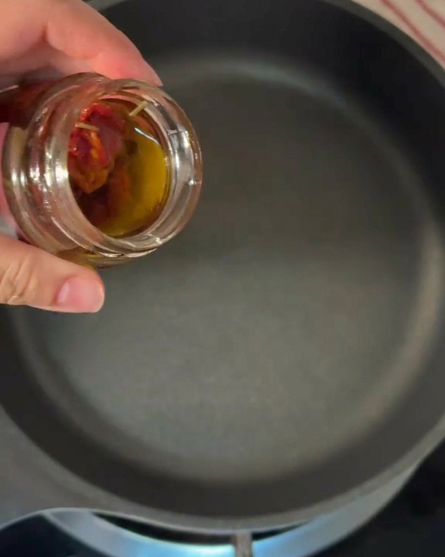 A hand tilting a small glass jar to pour orange-tinted infused olive oil into a heated black skillet.