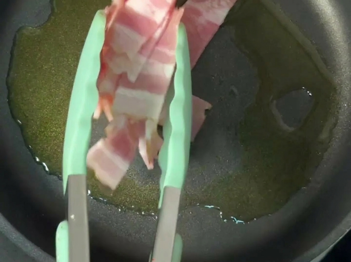 Crispy bacon strips frying in olive oil in a non-stick pan.