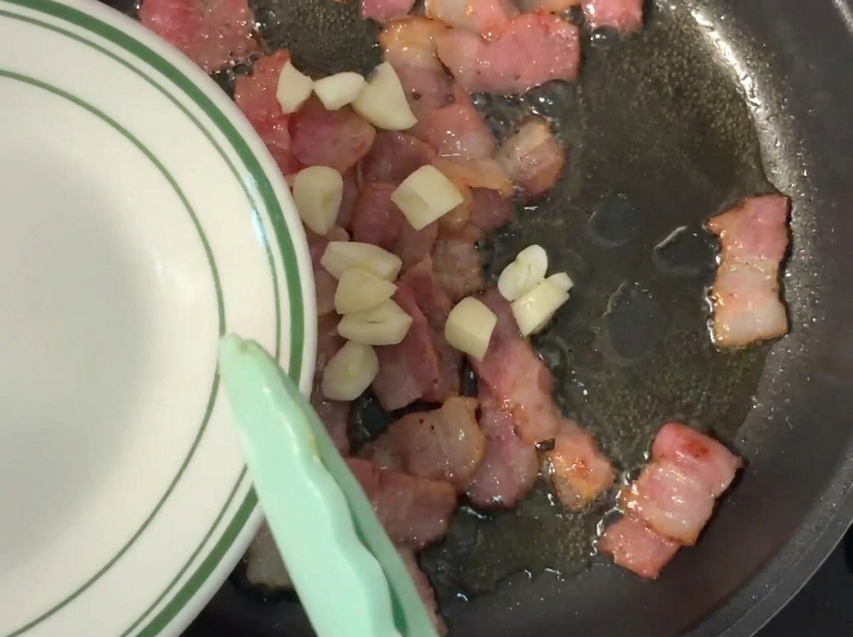 Freshly chopped garlic being pushed from a green-rimmed plate into a hot pan with sizzling, partially browned bacon pieces.