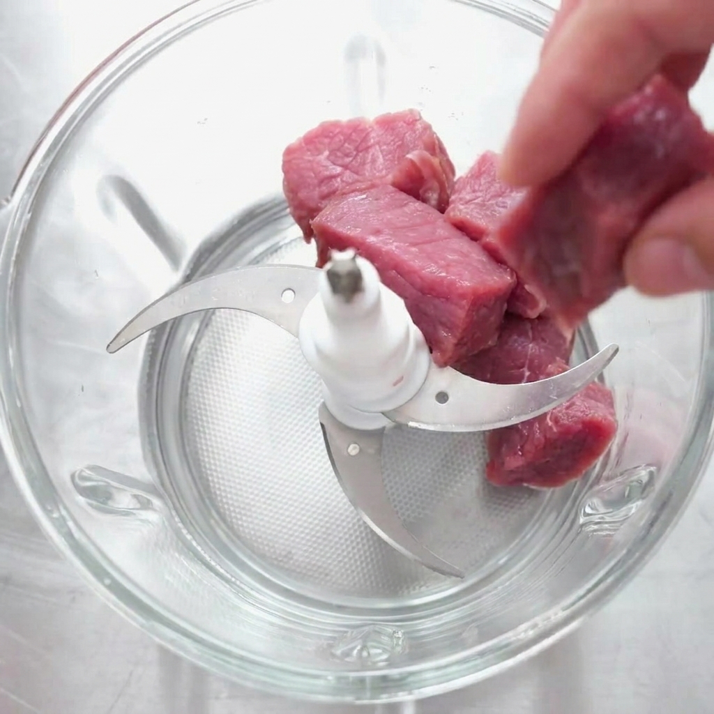Raw beef chunks being placed into a glass food chopper bowl equipped with a multi-blade attachment.