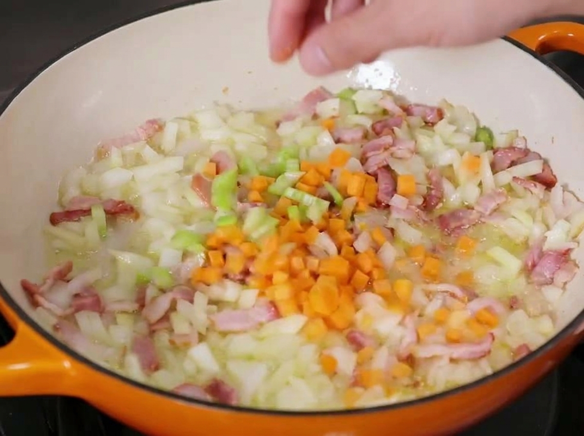 Diced carrots being sprinkled into a cast iron pan containing lightly sautéed onions, celery, and bacon pieces.