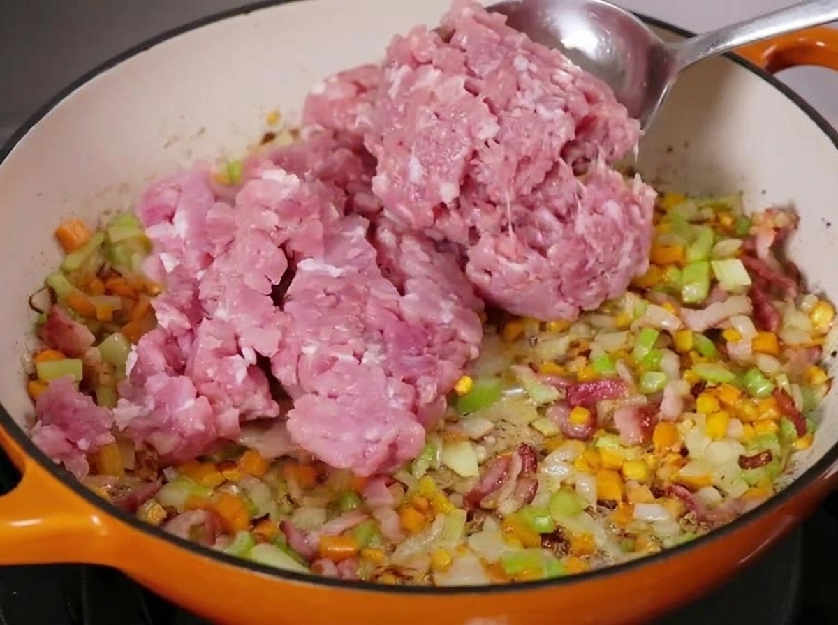 A large spoonful of raw ground meat being added to a simmering pan of diced vegetables and bacon.