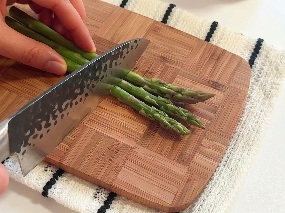 Hands using a chefs knife to slice fresh green asparagus stalks into short segments on a wooden cutting board.