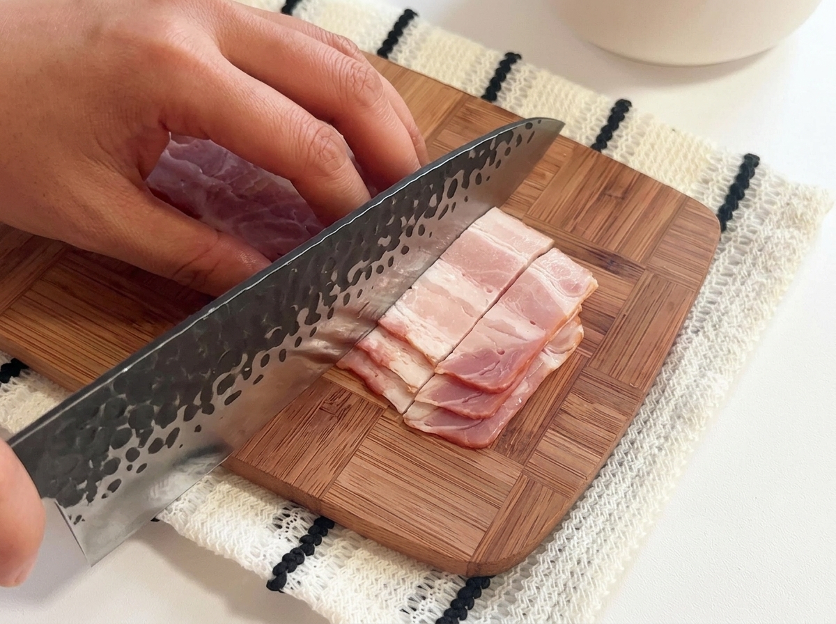 Raw bacon strips being cut into small pieces on a wooden cutting board with a large knife.