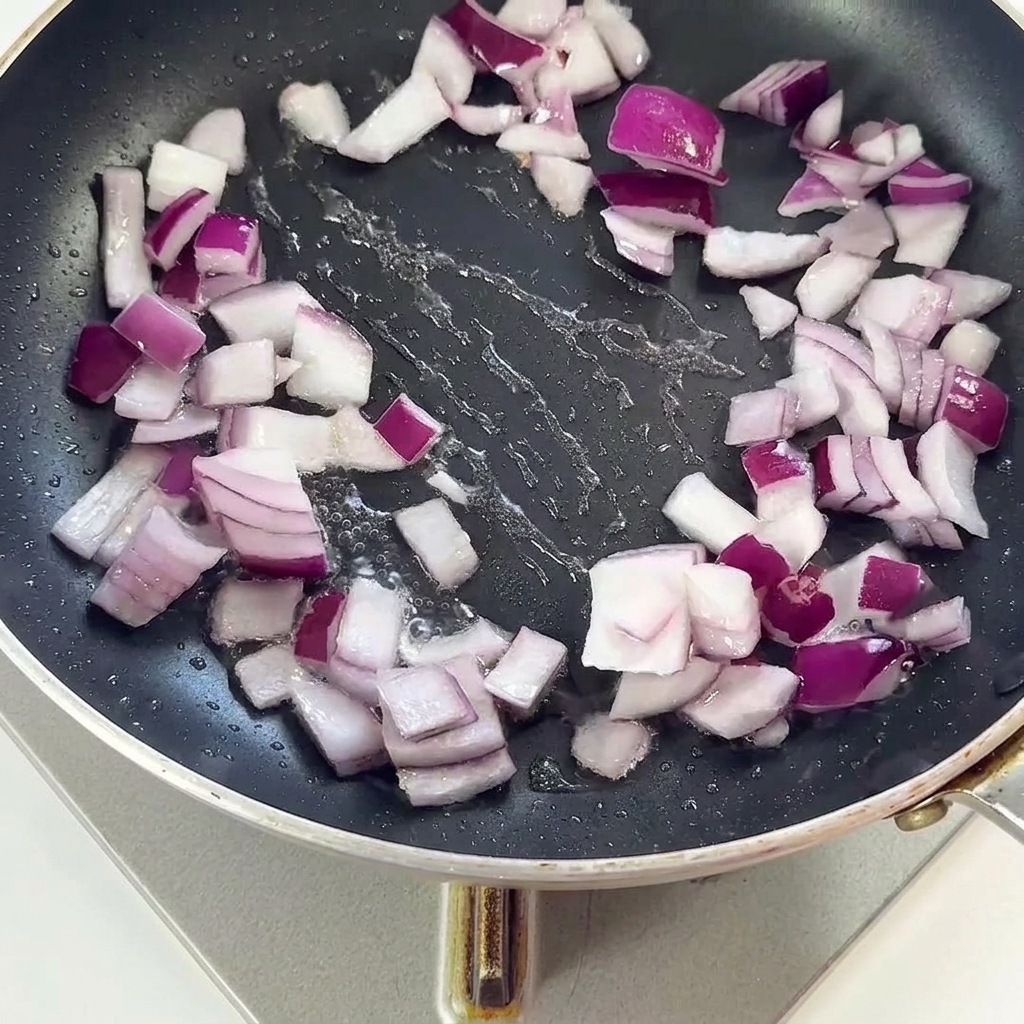 Bacon pieces and red onions stir-frying together in a black skillet.