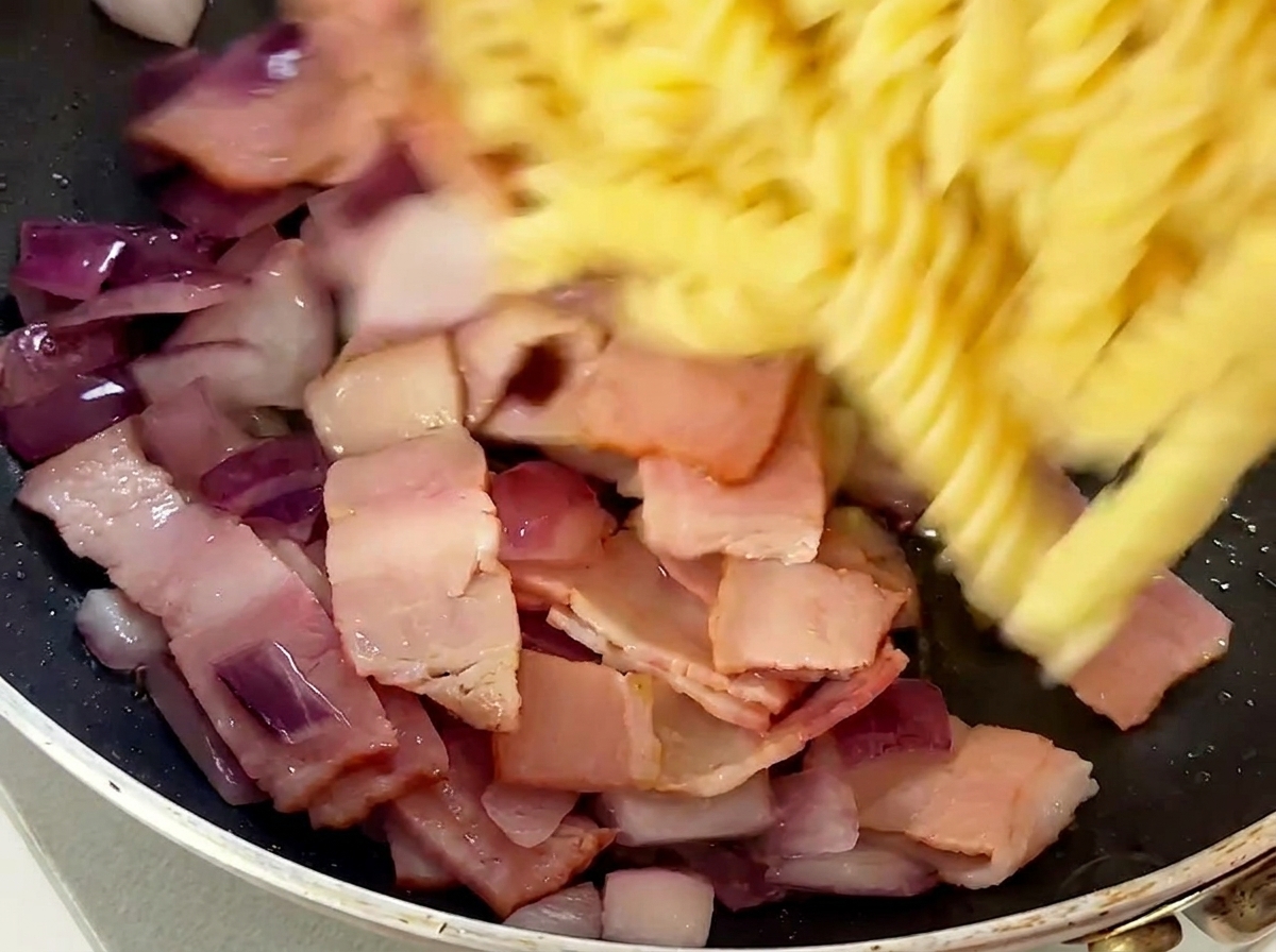 Cooked fusilli pasta being added to the skillet with bacon and onions.