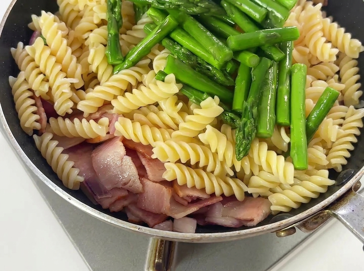 Asparagus spears and seasonings being tossed with pasta, bacon, and onions in a pan.