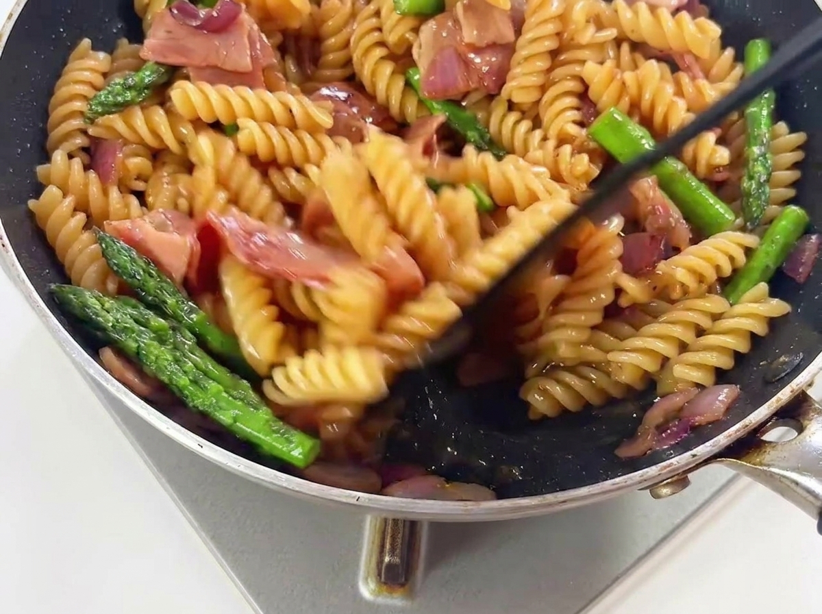 Pasta, green asparagus, and bacon pieces being tossed in a hot skillet to mix with the sauce.