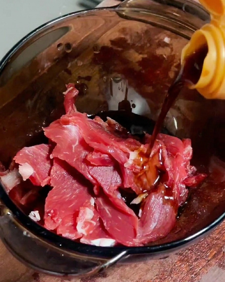 Soy sauce being poured onto fresh raw beef slices in a clear bowl.