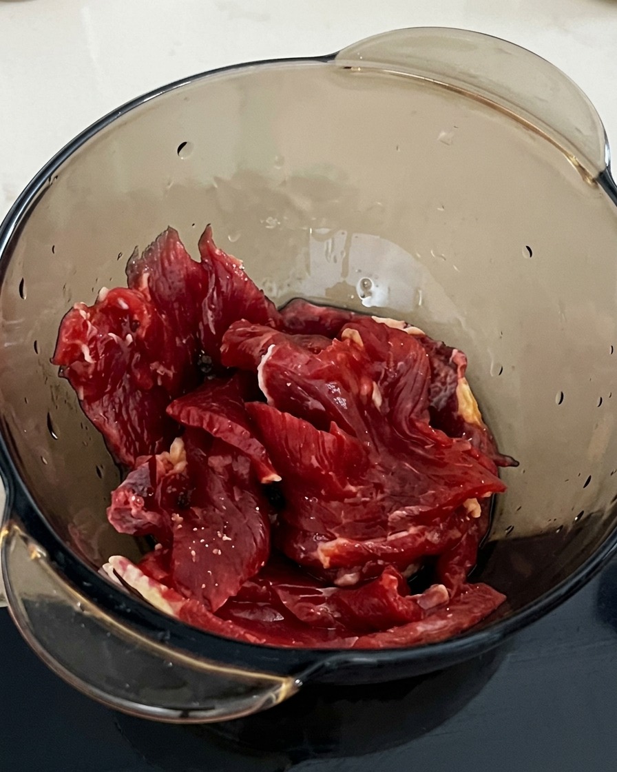 A spoonful of starch being added to a bowl containing beef slices and soy sauce.