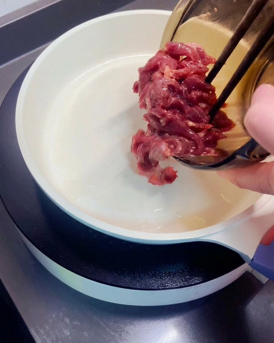 Raw marinated beef pieces being transferred from a glass bowl into a hot white skillet.