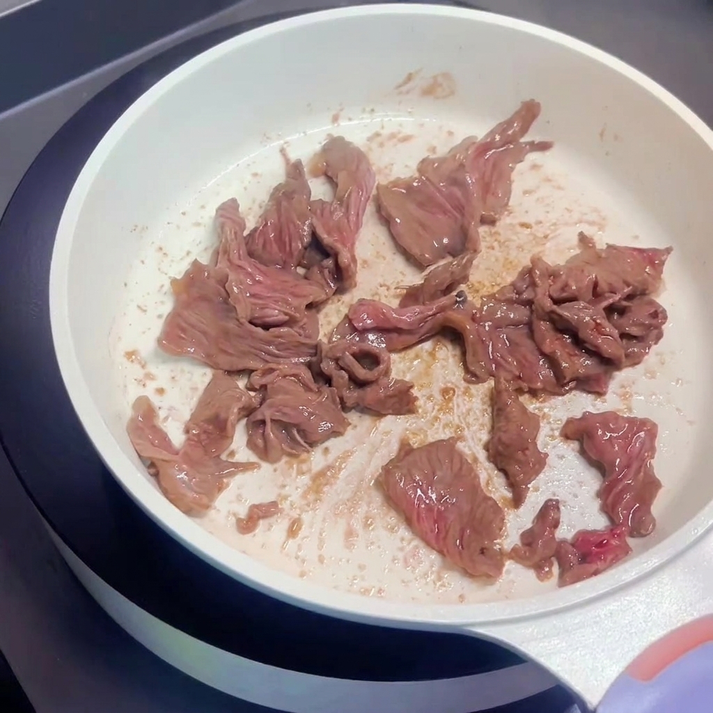 Beef slices being stir-fried in a white pan, showing browned surfaces.
