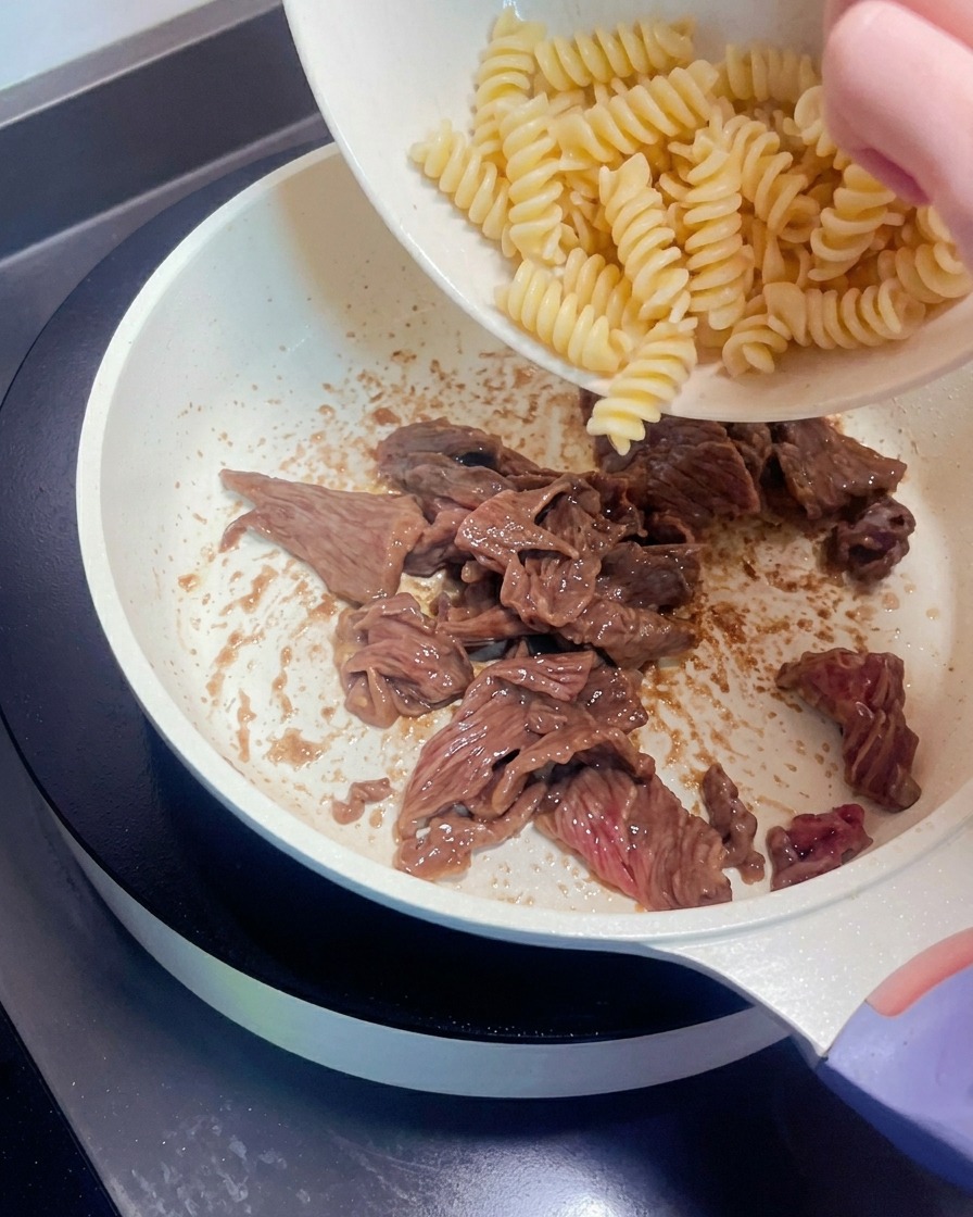 Spiral pasta being transferred from a bowl into a pan with cooked beef strips.