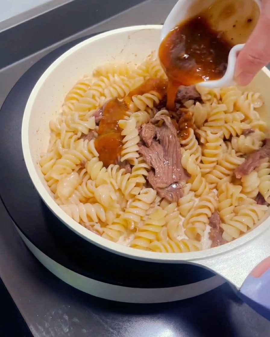 Pasta sauce being poured from a small dish over the beef and pasta in the pan.