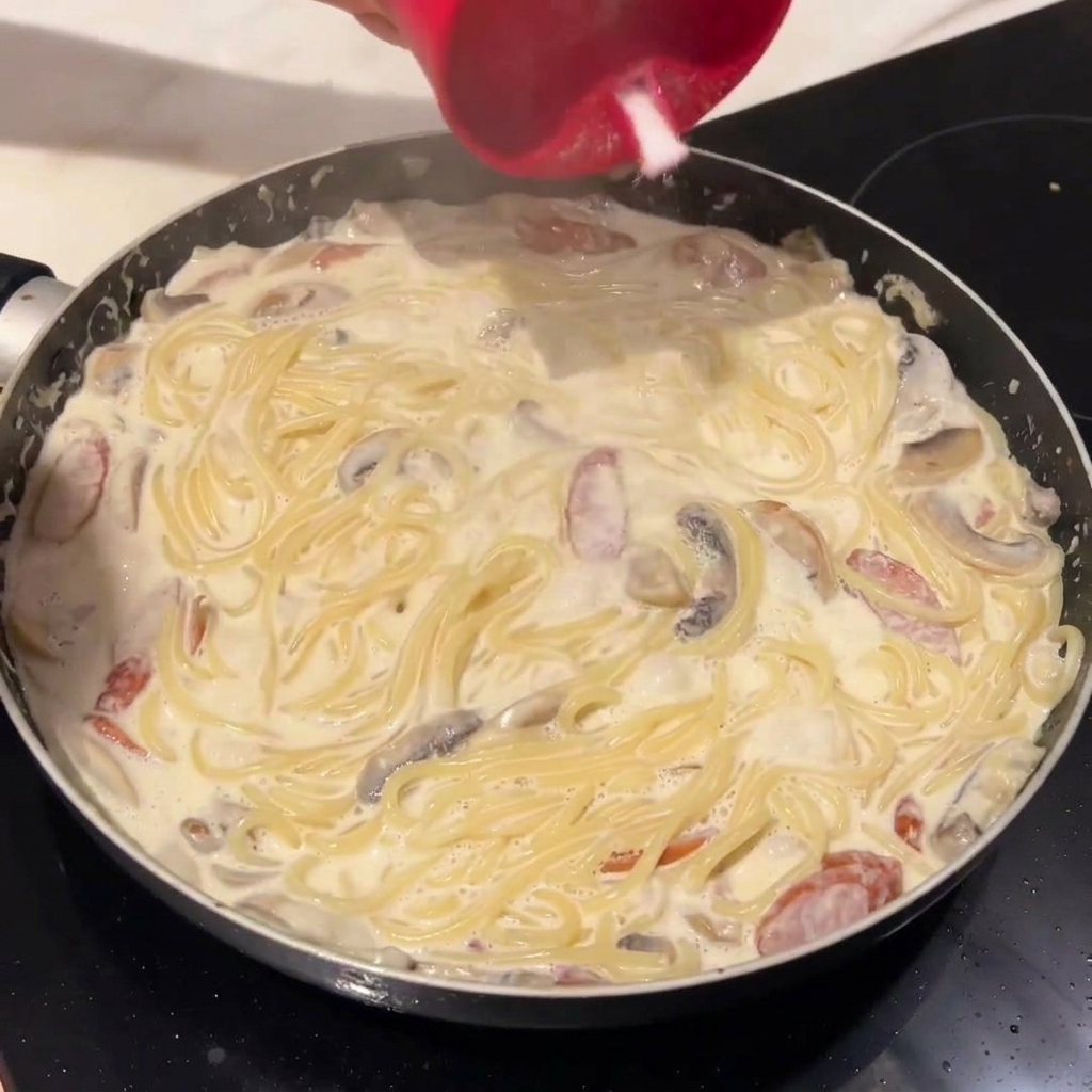 Cooked spaghetti being added to the creamy sauce in the pan.