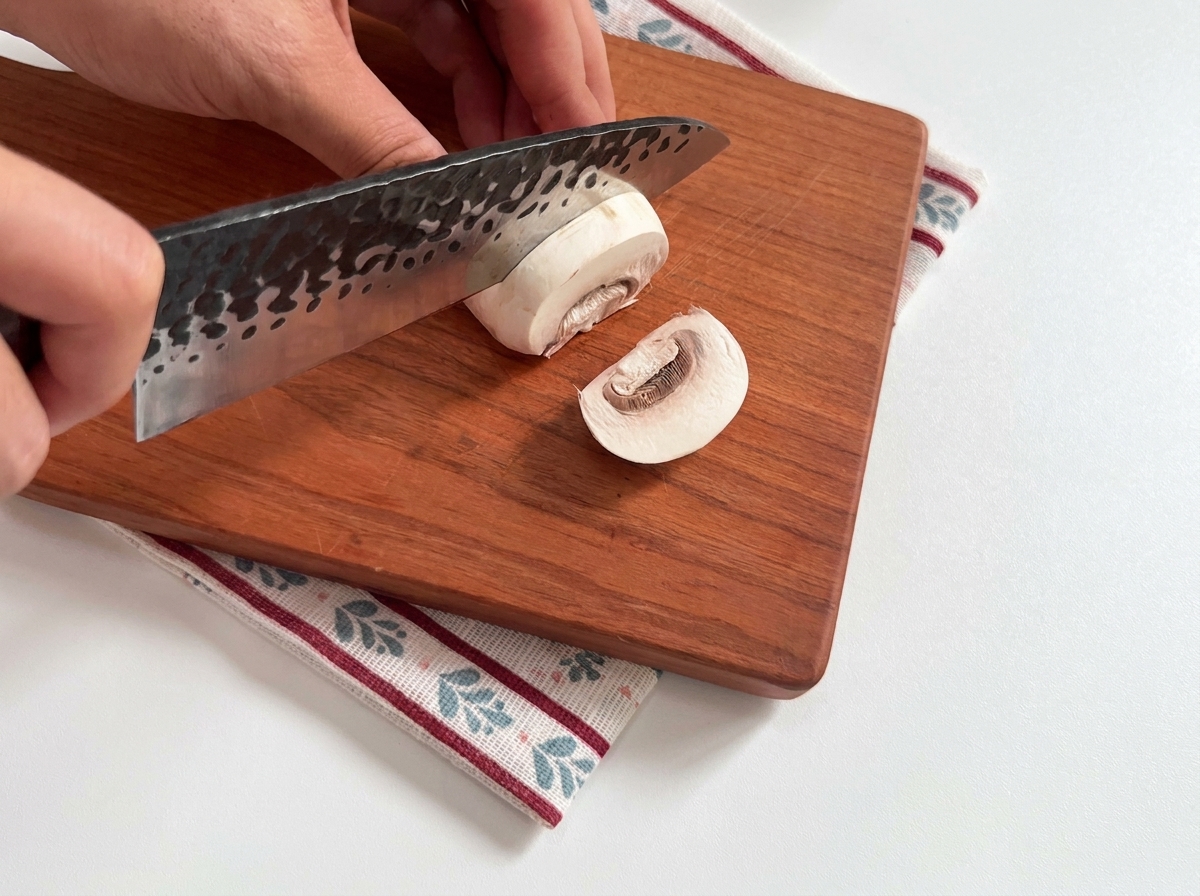 Fresh white button mushrooms being sliced on a wooden board.