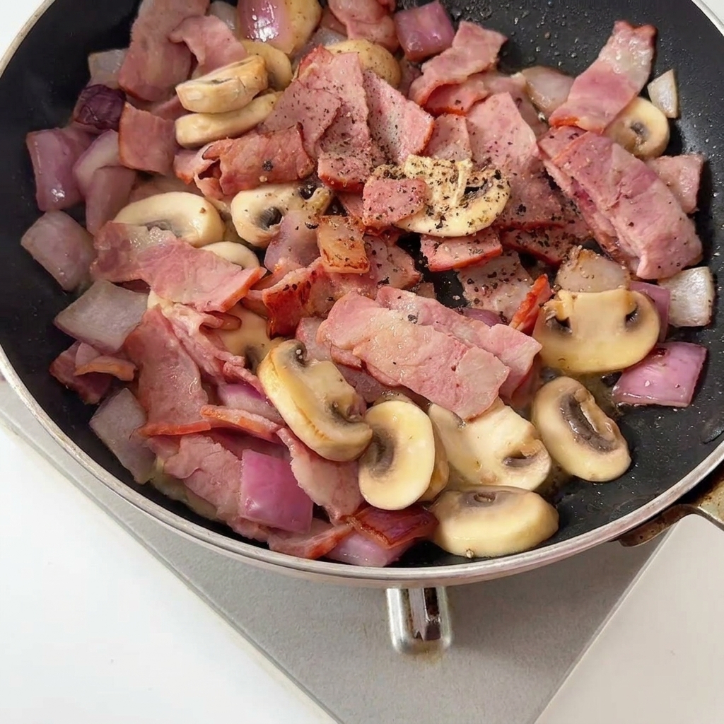 Freshly cracked black pepper being added over the sautéed bacon and mushroom mixture.