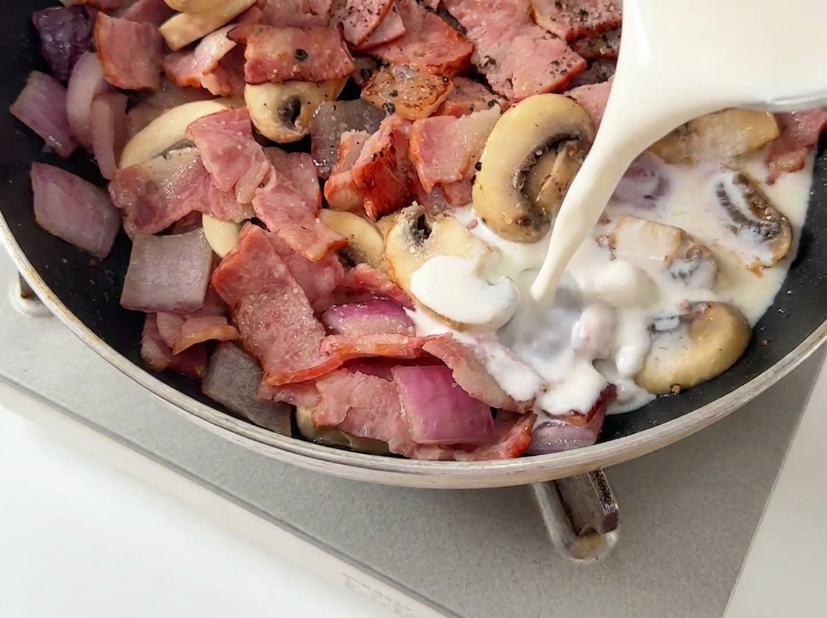 Milk and heavy cream being poured from a glass measuring cup into a pan containing bacon and mushrooms.