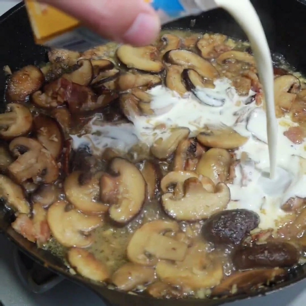 Heavy cream being poured into a pan containing sautéed mushrooms, bacon, and golden-brown onions.