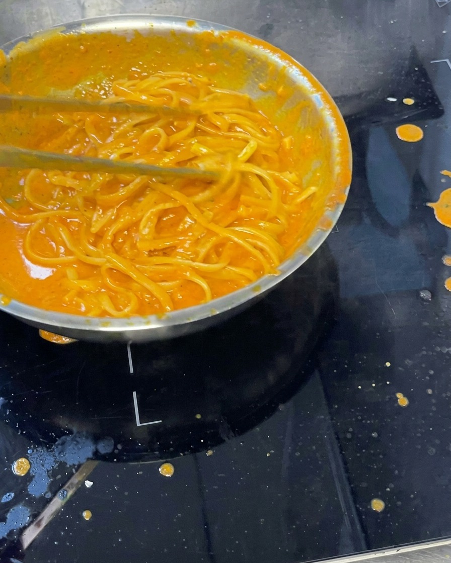 Creamy pasta mixed with roasted pepper sauce in a pan, with a ladle mixing the ingredients.