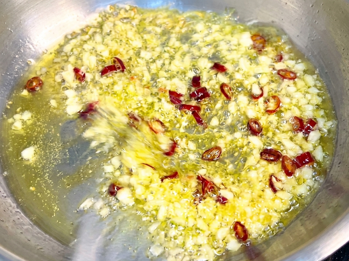 Pasta being tossed in a pan with golden garlic and chili flakes, coating in a glossy emulsified sauce.