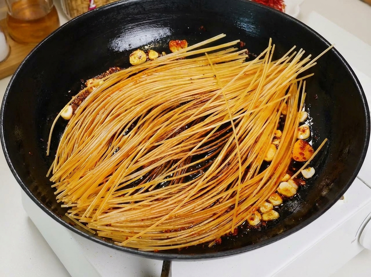 A ladle pouring red tomato broth onto pan-seared spaghetti in a black pan.
