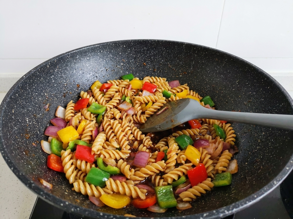 Rotini pasta, beef strips, and colorful bell peppers being stir-fried with a gray spatula in a wok, thoroughly coated in a glossy dark sauce.