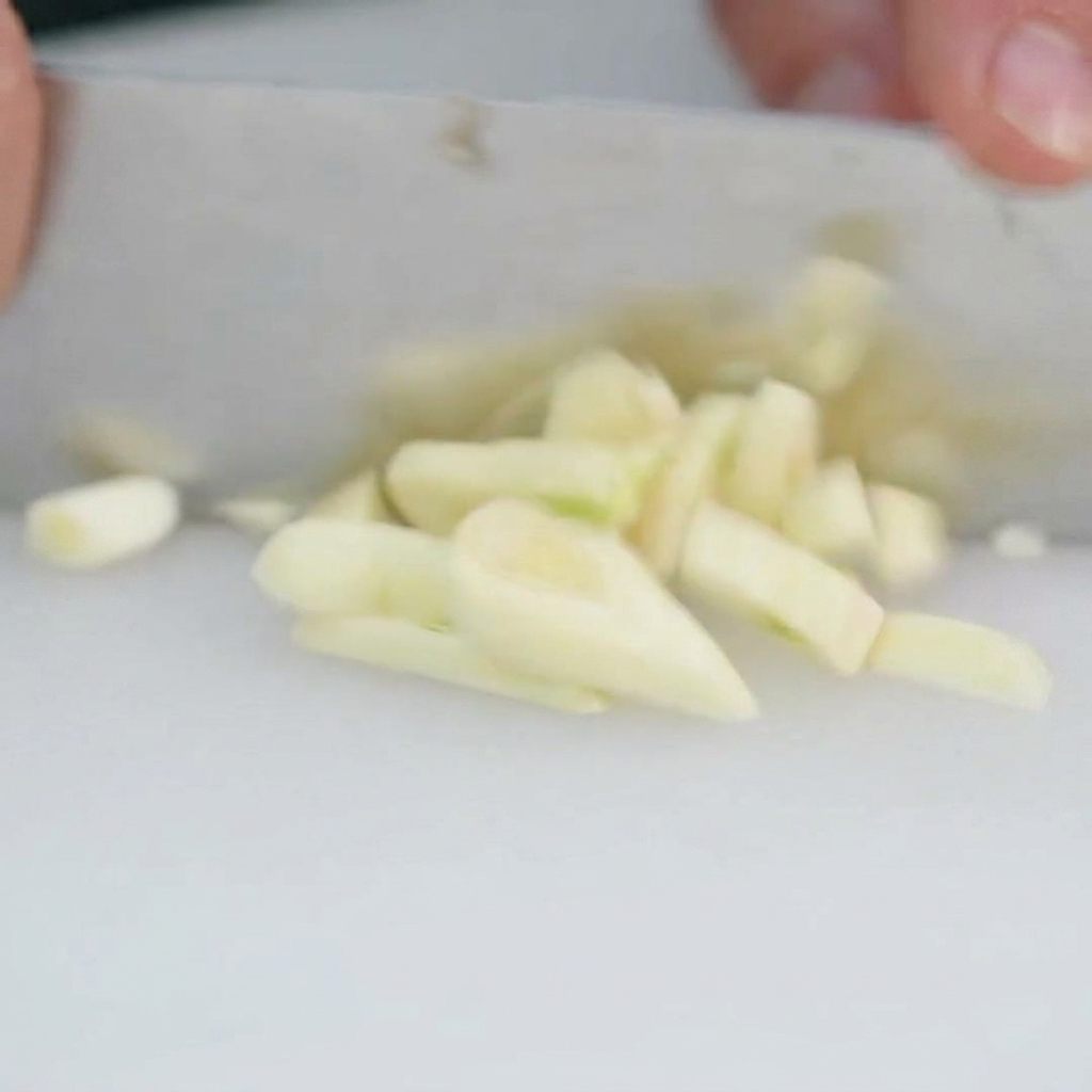 Fresh garlic cloves being finely minced into small pieces on a cutting board.