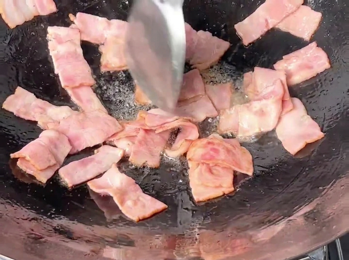 Pink rectangular bacon pieces being stir-fried in a dark iron wok using a metal spatula.