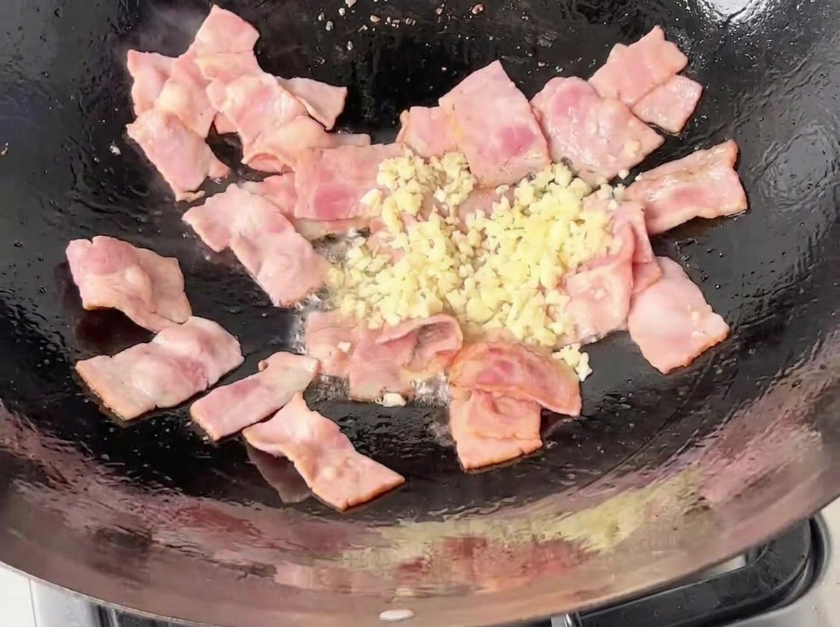 Finely minced white garlic being added to the pan with sizzling, partially cooked bacon pieces.