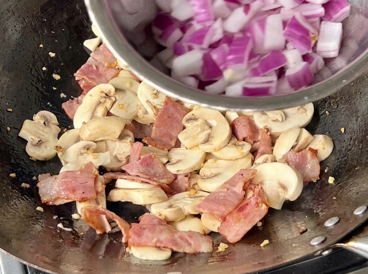 A pan filled with sliced mushrooms, bacon bits, and freshly added diced purple onions being stir-fried together.