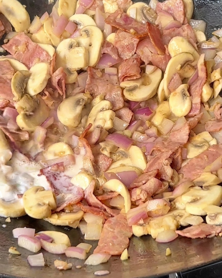 Milk being poured into a pan containing fried bacon, sautéed mushrooms, and onions.